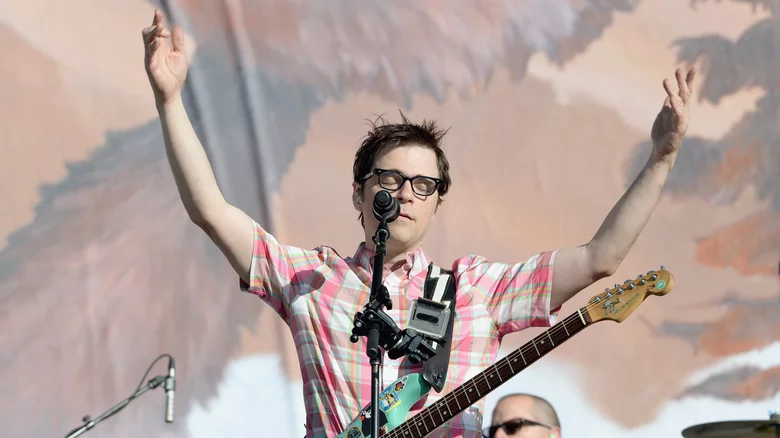 A photo of musician Rivers Cuomo during a performance with his eyes closed and his arms up.